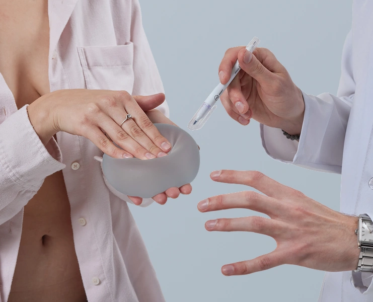 A surgeon holding a surgical marker and pointing while a patient examines a silicone breast implant during a reconstructive surgery consultation.