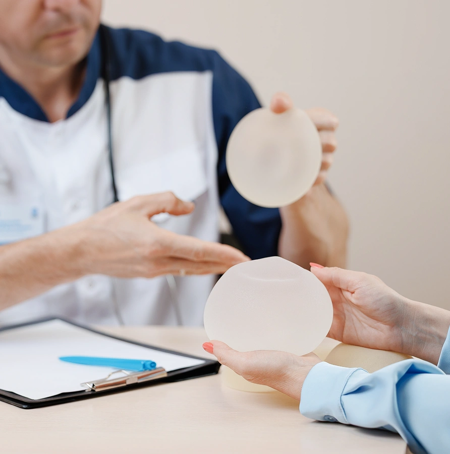 Plastic surgeon holding a silicone breast implant while a patient examines another implant during a consultation.