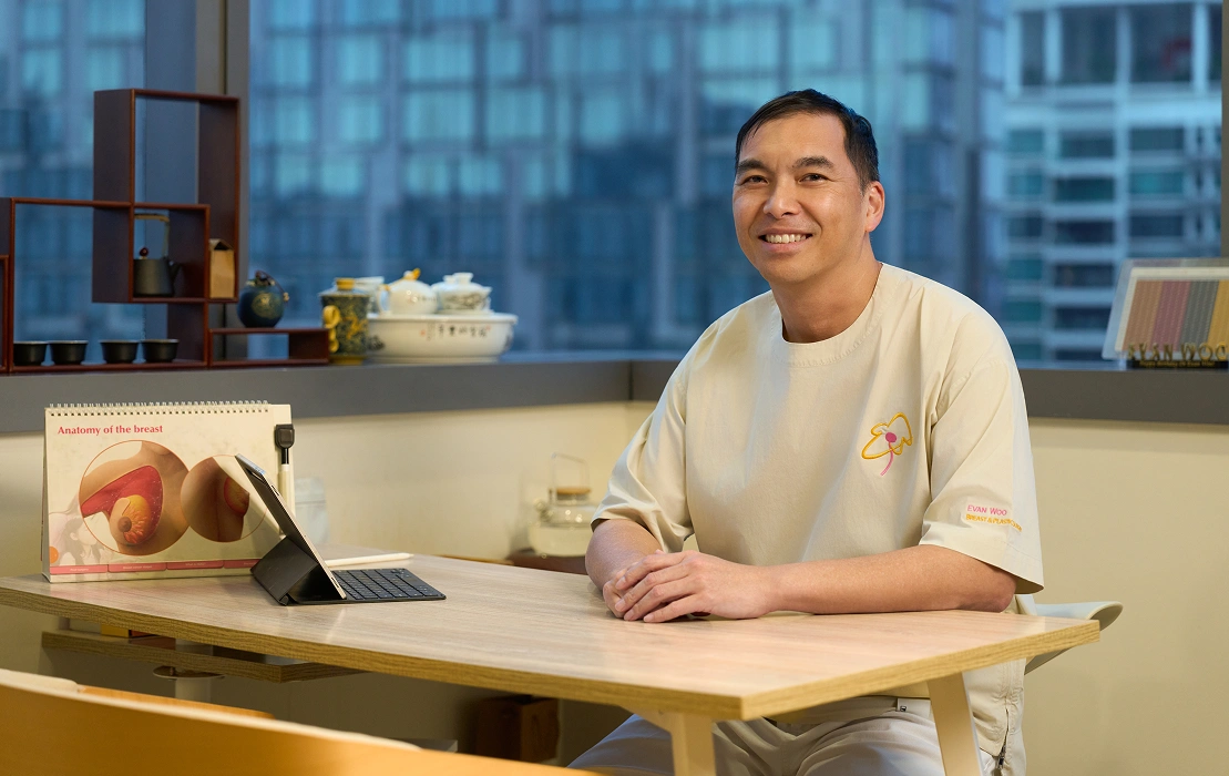 Dr Evan Woo smiling at his clinic consultation desk beside a breast anatomy educational display.