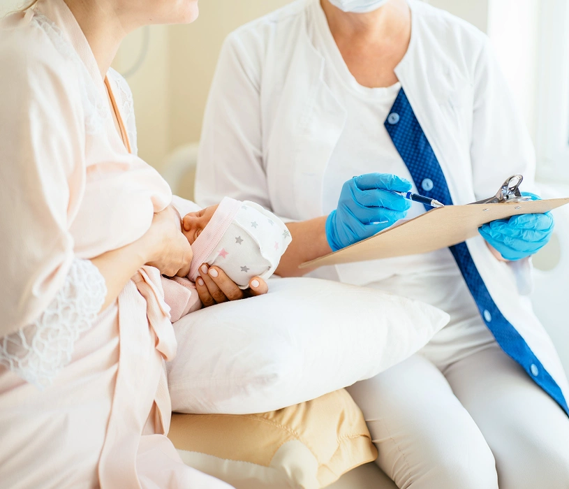 A mother breastfeeding her newborn baby while a healthcare provider in blue gloves writes on a clipboard.