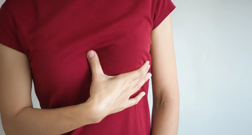 A woman in a red t-shirt holding her hand over her chest indicating breast pain or screening awareness.