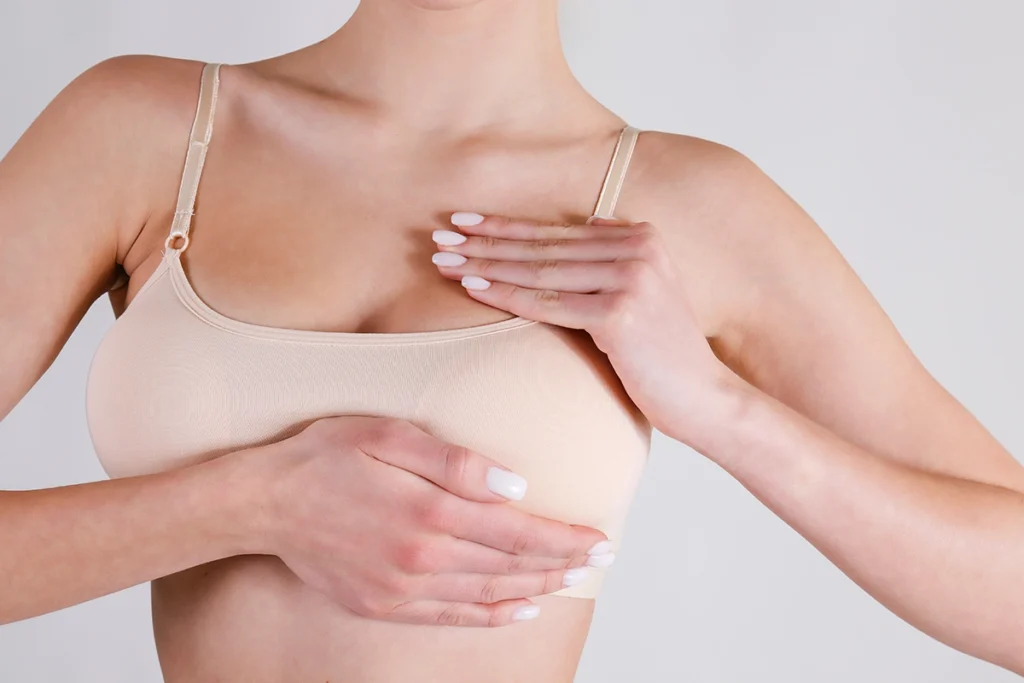 A woman in a beige camisole placing both hands on her chest to perform a routine breast self-examination.